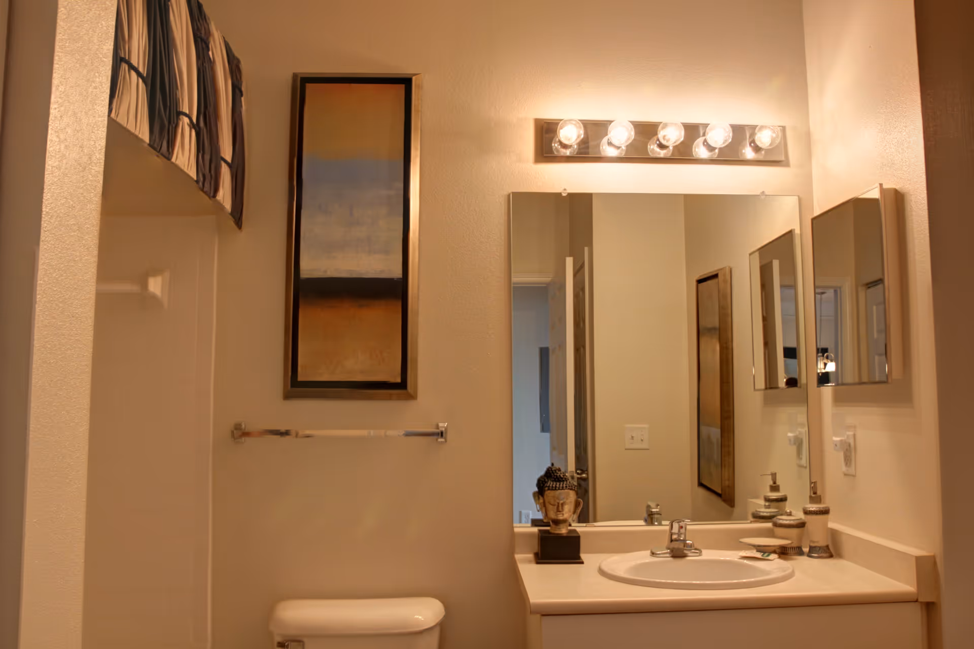 A bathroom interior featuring a white sink with a countertop, a large mirror above the sink, and a light fixture with six bulbs. To the left, there is a toilet and a towel rack mounted on the wall. Above the towel rack, a framed abstract painting is hung. The shower area is partially visible with a curtain rod and a patterned shower curtain.