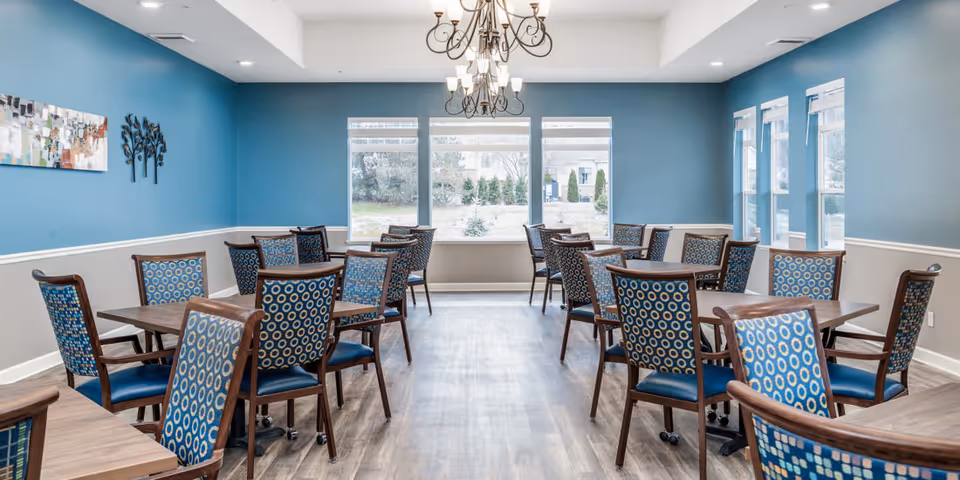 A dining room with multiple wooden tables and blue patterned chairs arranged neatly. The walls are painted blue with white trim, and there are large windows letting in natural light. Two chandeliers hang from the ceiling, and decorative wall art is visible on the left wall.