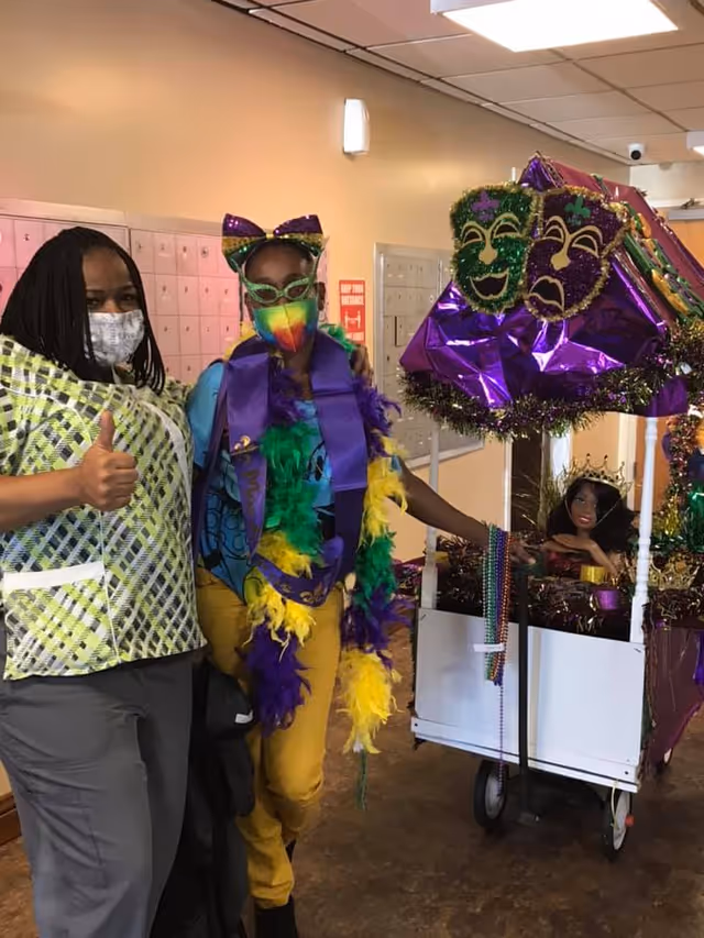 Two women standing indoors near a decorated cart with Mardi Gras themed masks and decorations. One woman is wearing a patterned shirt and a face mask, giving a thumbs up. The other woman is dressed in colorful Mardi Gras attire including a rainbow face mask, green glasses, a purple sash, and feather boas in green, yellow, and purple. The cart beside them is adorned with purple, green, and gold decorations and beads, and contains a doll wearing a crown.