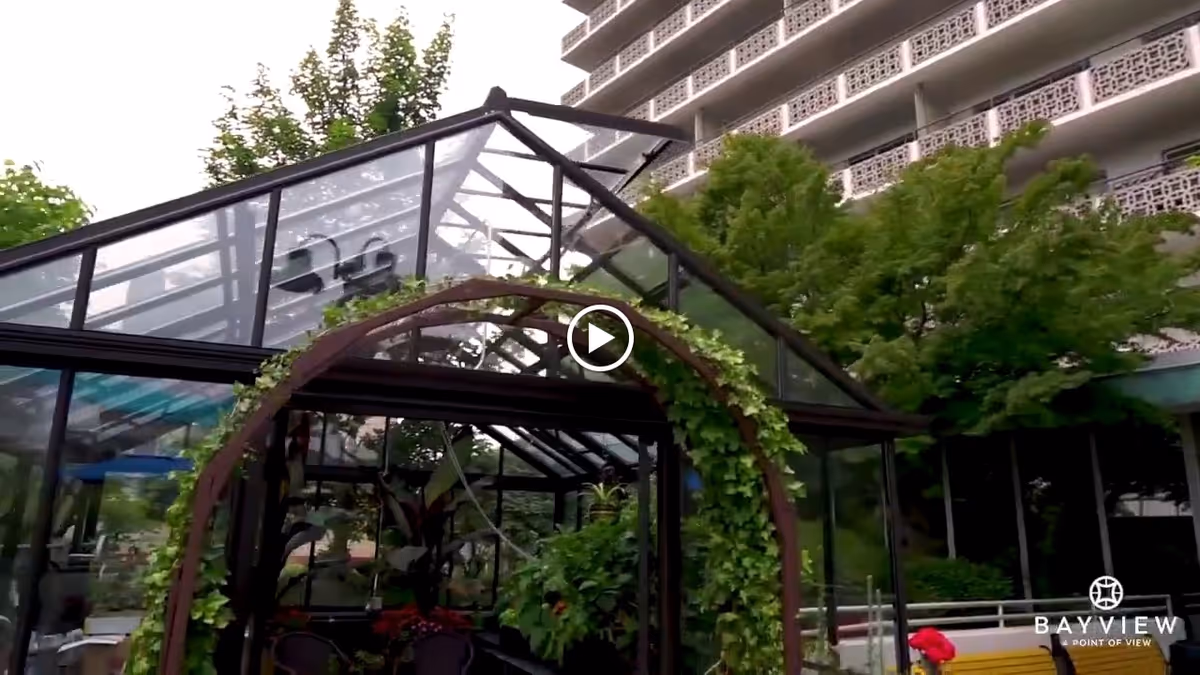 Glass greenhouse structure with plants inside and a metal arch covered in green vines in front. Behind the greenhouse, there is a multi-story building with decorative balcony railings and trees surrounding the area.