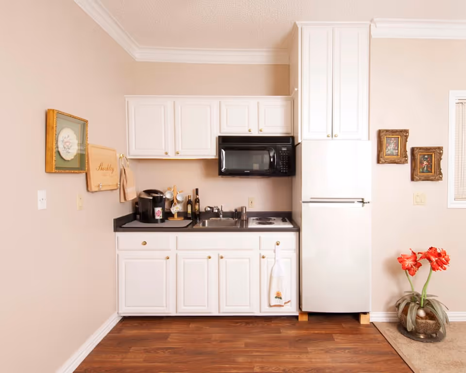 A small kitchen area with white cabinets, a black countertop, a black microwave mounted above a small electric stove, a white refrigerator, a coffee maker, and a sink. The walls are beige with framed artwork and a wooden cutting board hanging. There is a vase with red flowers on the floor to the right.