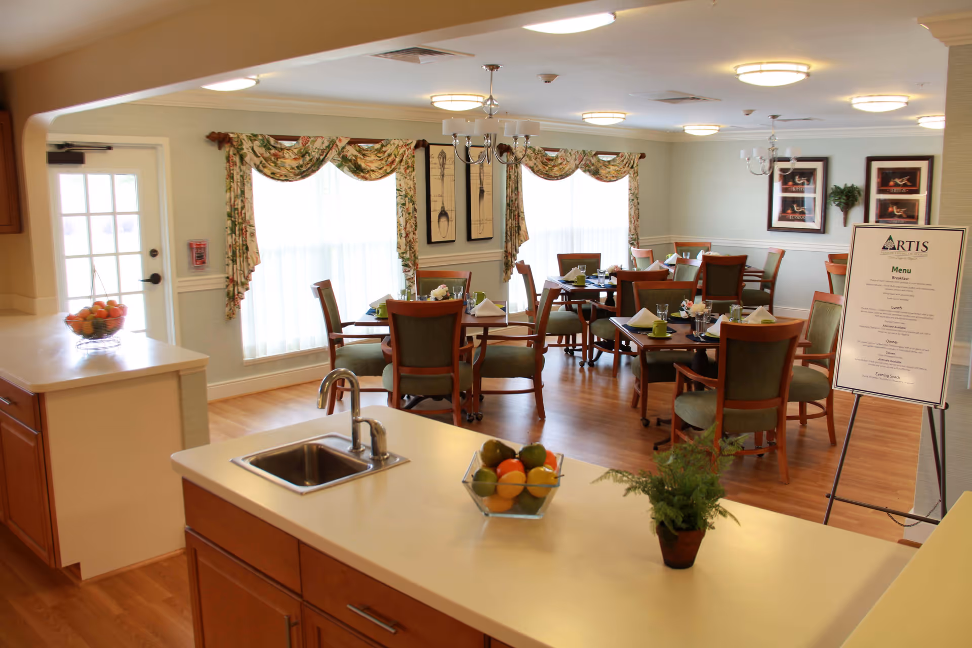 A bright dining room in a senior living facility with several wooden tables and chairs arranged neatly. The room has large windows with floral curtains allowing natural light to fill the space. A kitchen counter with a small sink, a bowl of fruit, and a potted plant is visible in the foreground. A menu board labeled 'ARTIS' stands near the dining area.