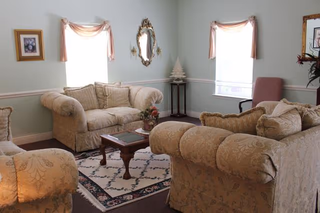 A cozy living room with three beige patterned sofas arranged around a wooden coffee table on a decorative rug. The room has light green walls, two windows with sheer curtains and pink valances, a small side table with a white decorative tree, a wall mirror, and framed artwork.