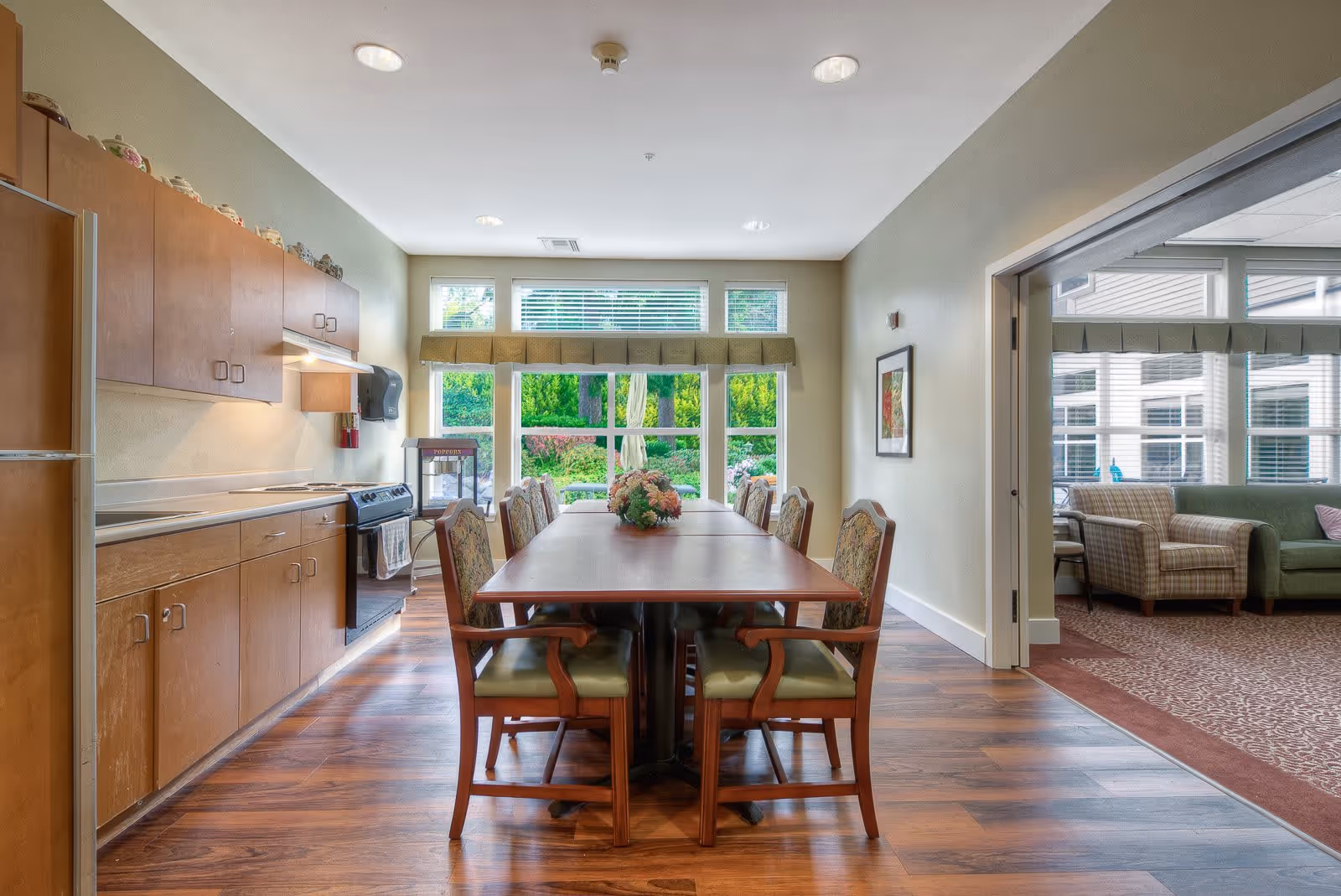 A bright dining area in a senior living facility with a long wooden table surrounded by eight cushioned chairs. To the left is a kitchen area with wooden cabinets, a stove, and a refrigerator. Large windows at the end of the room let in natural light and offer a view of greenery outside. To the right, an open doorway leads to a living room with armchairs and a sofa.