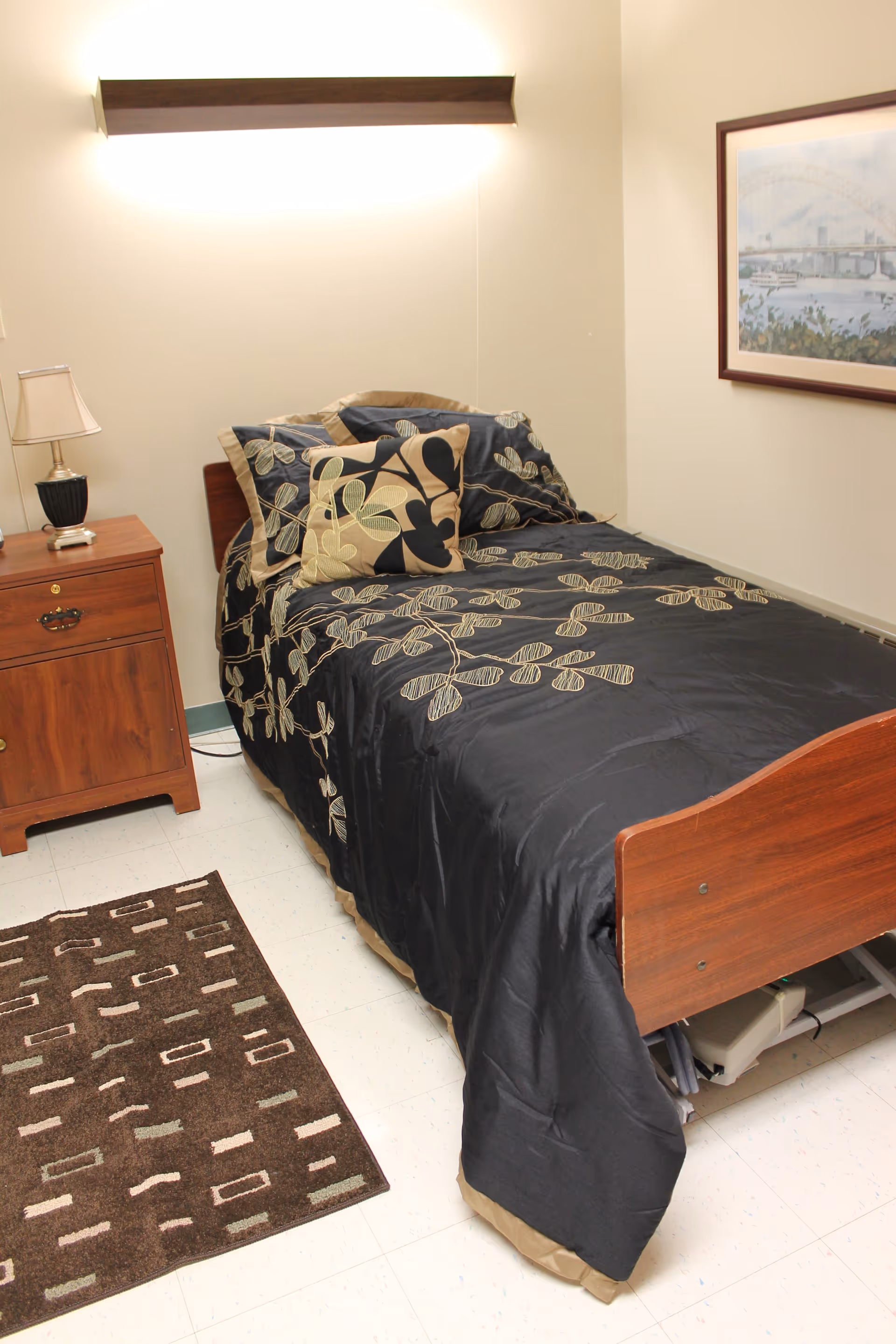 A single bed with a dark floral patterned bedspread and matching pillows in a small room. Next to the bed is a wooden nightstand with a lamp and a small item on it. A framed picture hangs on the wall above the bed. There is a patterned rug on the floor beside the bed.