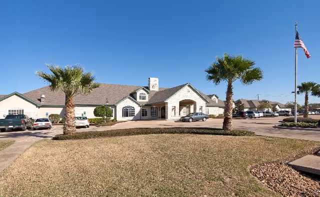 Exterior view of Carriage Inn Lake Jackson facility showing a single-story building with a light-colored stone facade, a driveway with parked cars, palm trees, and an American flag on a flagpole under a clear blue sky.