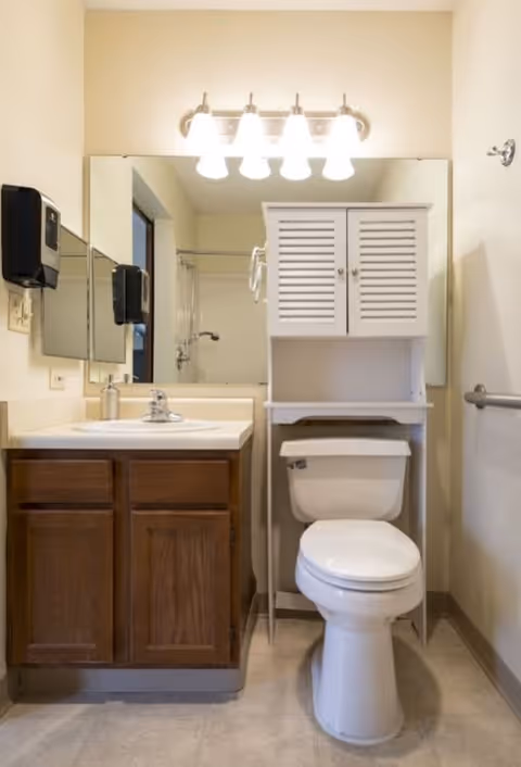 A clean bathroom featuring a white toilet with a white storage cabinet above it, a wooden vanity with a white sink and silver faucet, a large mirror above the sink, and a four-light fixture mounted on the wall. There is a soap dispenser mounted on the wall next to the mirror and a grab bar on the right wall.