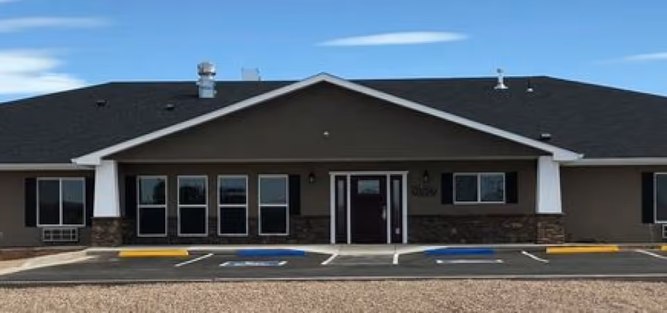 Front exterior view of a single-story assisted living facility building with a dark roof, multiple windows, a central entrance door, and a parking area with designated handicap parking spaces in front.