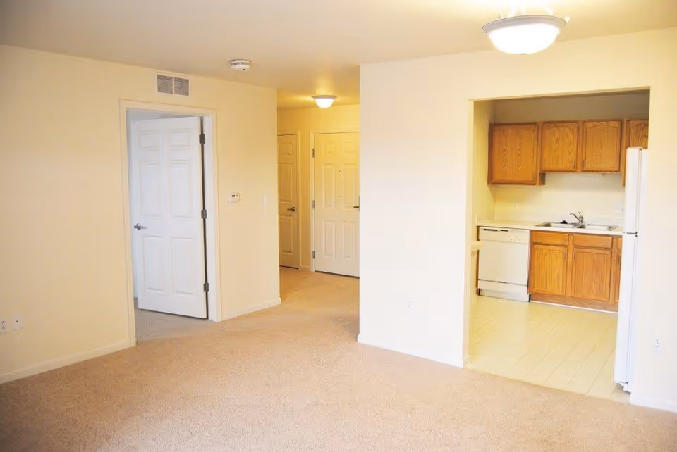 Interior view of an apartment with beige carpeted flooring and cream-colored walls. There is an open doorway leading to a small kitchen area with wooden cabinets, a white dishwasher, a white refrigerator, and a sink. Another open door leads to a separate room, and a hallway with a closed door is visible in the background. Ceiling lights illuminate the space.