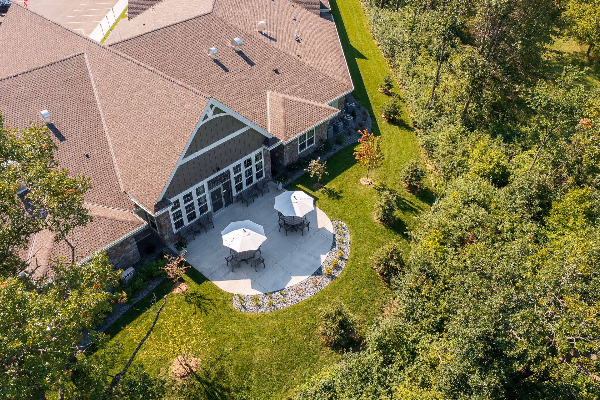 Aerial view of an outdoor patio area at NorBella Senior Living - Champlin, featuring two tables with umbrellas and chairs on a concrete patio surrounded by green grass and trees, adjacent to a building with a brown roof.