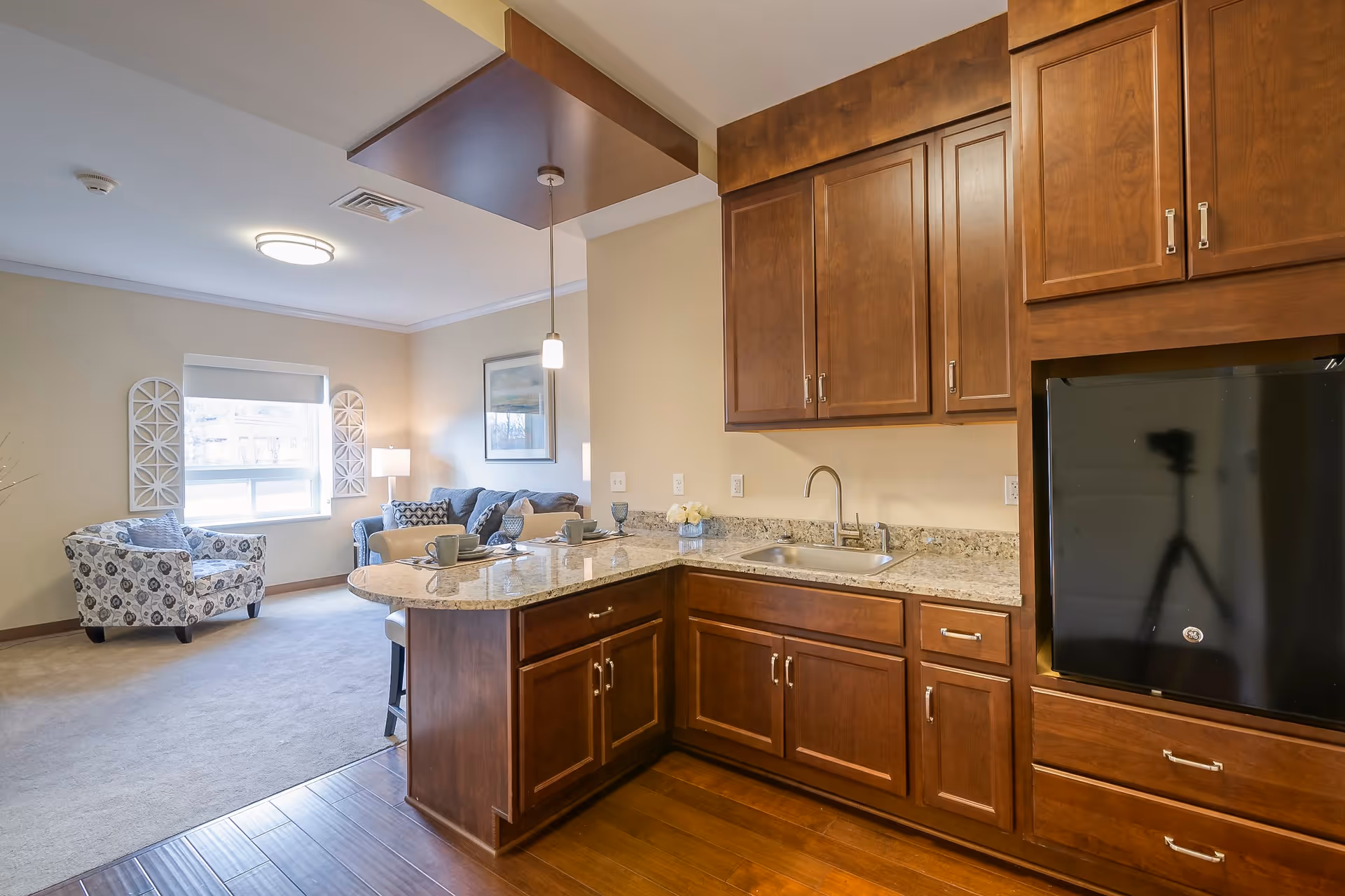 Interior view of a senior living facility showing a kitchen area with wooden cabinets, granite countertops, a sink, and a small black refrigerator. Adjacent to the kitchen is a living room area with a patterned armchair, a sofa, a window with blinds, and wall decorations.