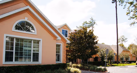 Exterior view of a peach-colored building with large windows and a gabled roof, surrounded by trees and landscaping under a partly cloudy sky.
