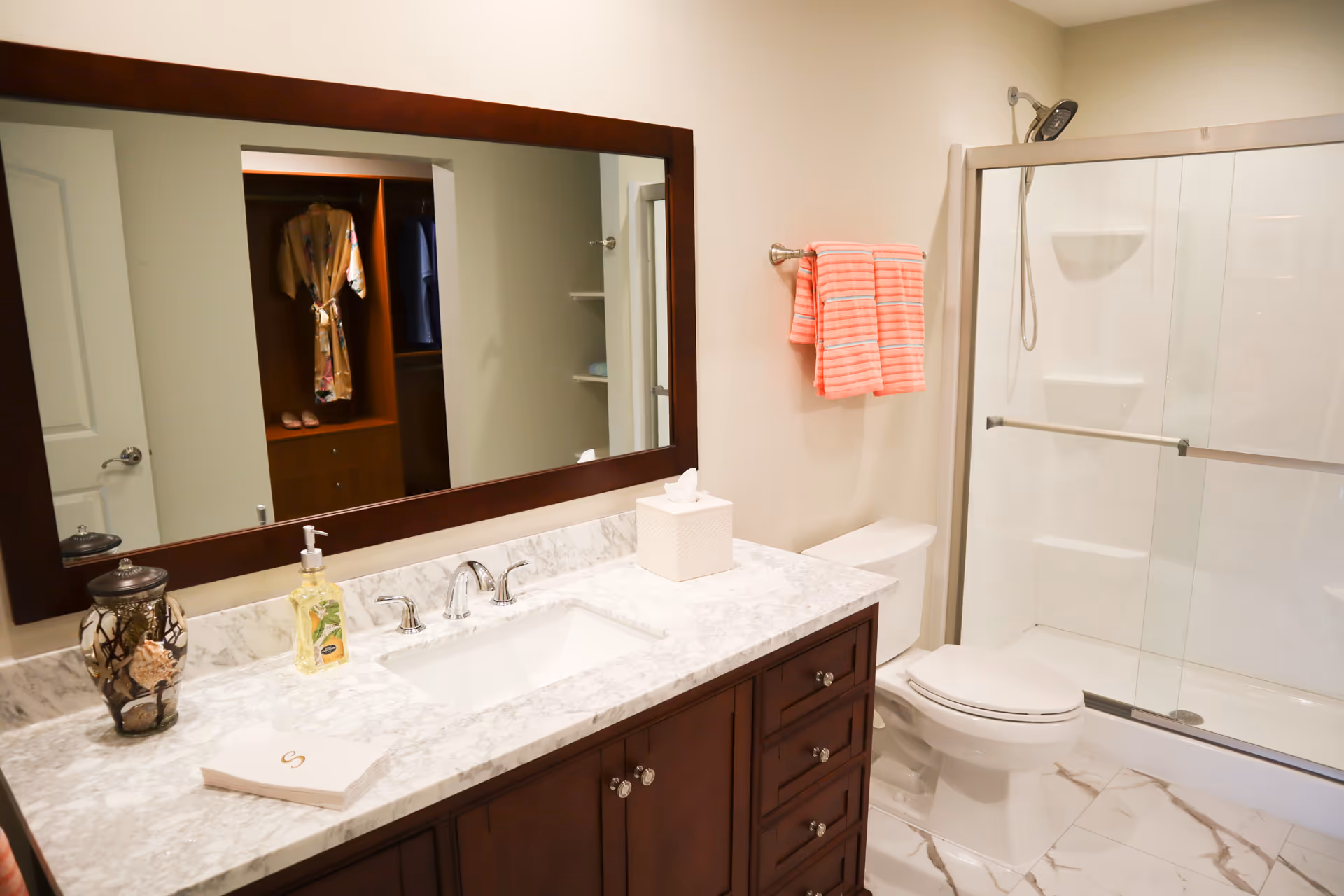 A clean and modern bathroom with a marble countertop vanity featuring a sink, soap dispenser, tissue box, and decorative jar. Above the vanity is a large mirror reflecting a walk-in closet with hanging clothes. To the right is a toilet and a glass-enclosed shower with a chrome showerhead. A towel rack with two pink striped towels is mounted on the wall above the toilet.