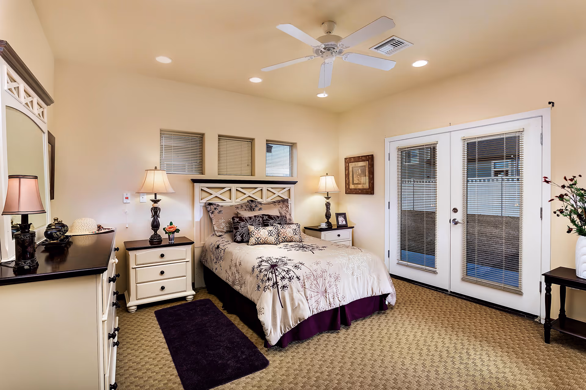 A cozy senior living bedroom with a bed featuring floral bedding and multiple pillows, two nightstands with lamps on either side, a dresser with a mirror, a ceiling fan, and French doors with blinds leading outside. The room has beige walls and carpeted flooring.