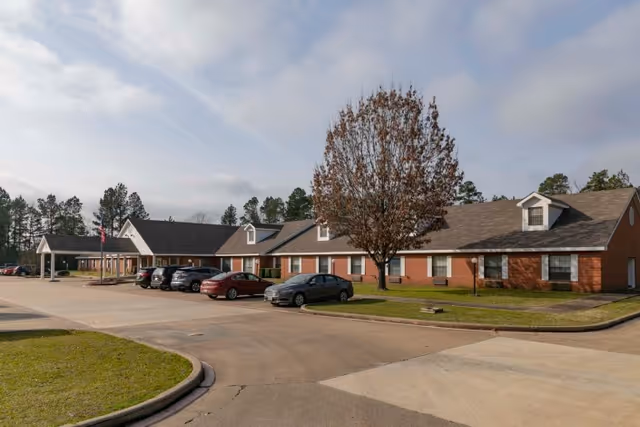Exterior view of Legend Oaks Healthcare And Rehabilitation Center - Gladewater, showing a single-story brick building with a pitched roof, several parked cars, a tree with brown leaves, and a partly cloudy sky.