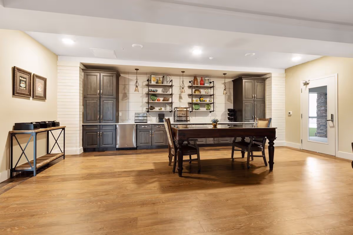 Communal dining area with a wooden table and chairs in front of a built-in kitchenette with cabinets and open shelving.