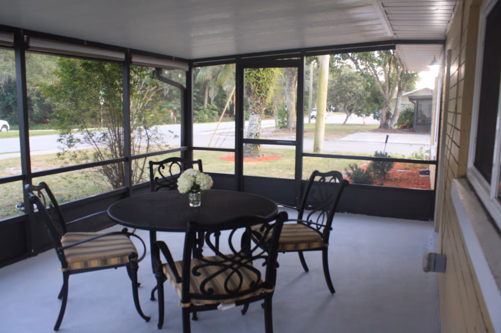 A screened-in porch area with a round black metal table and four matching chairs with striped cushions. A small vase with white flowers is placed on the table. Outside the screened porch, there is a view of a lawn, trees, and a street.
