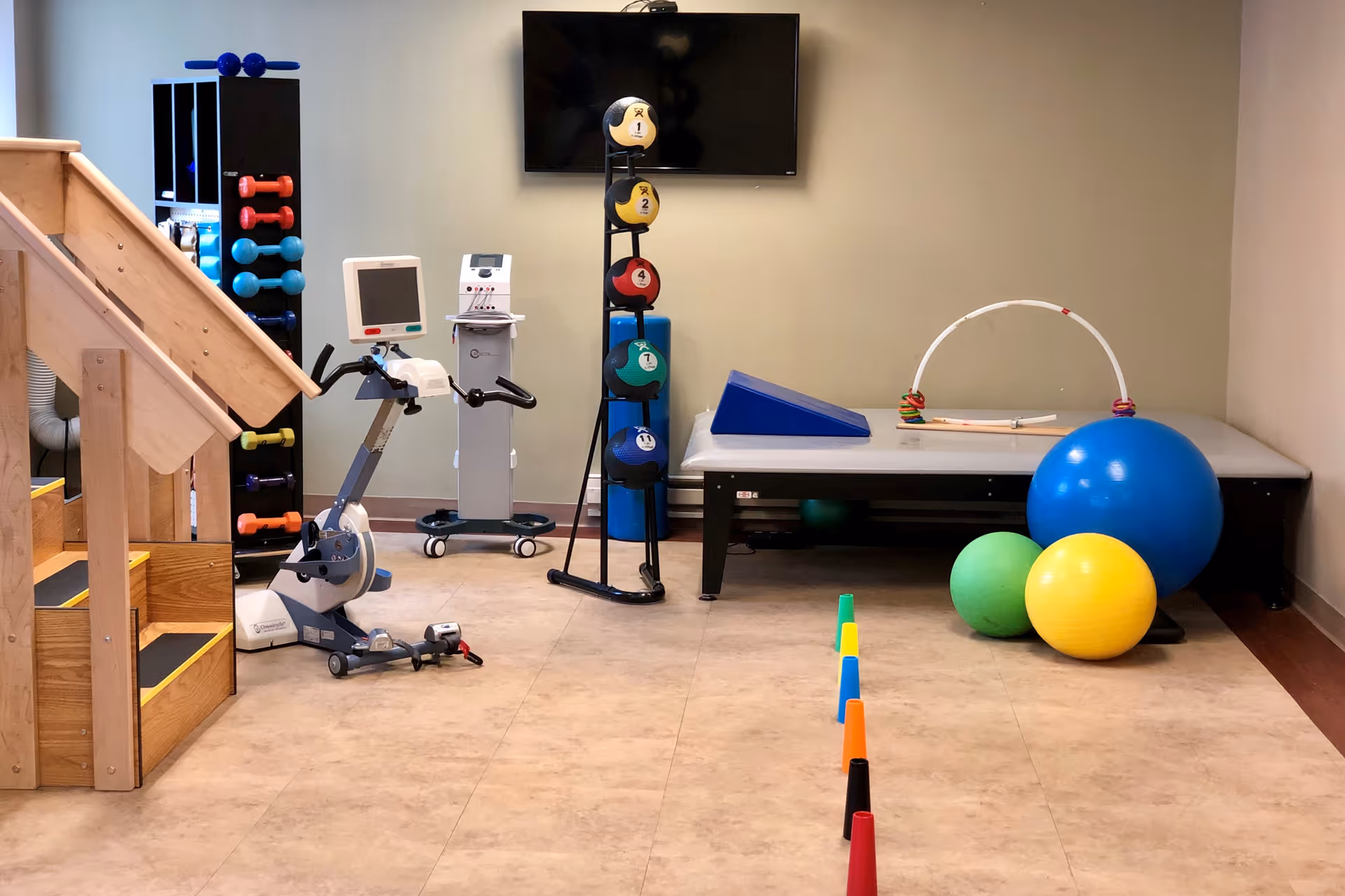 A therapy room featuring various exercise equipment, including a stationary bike, weights, colorful exercise balls, and a set of stairs for rehabilitation.
