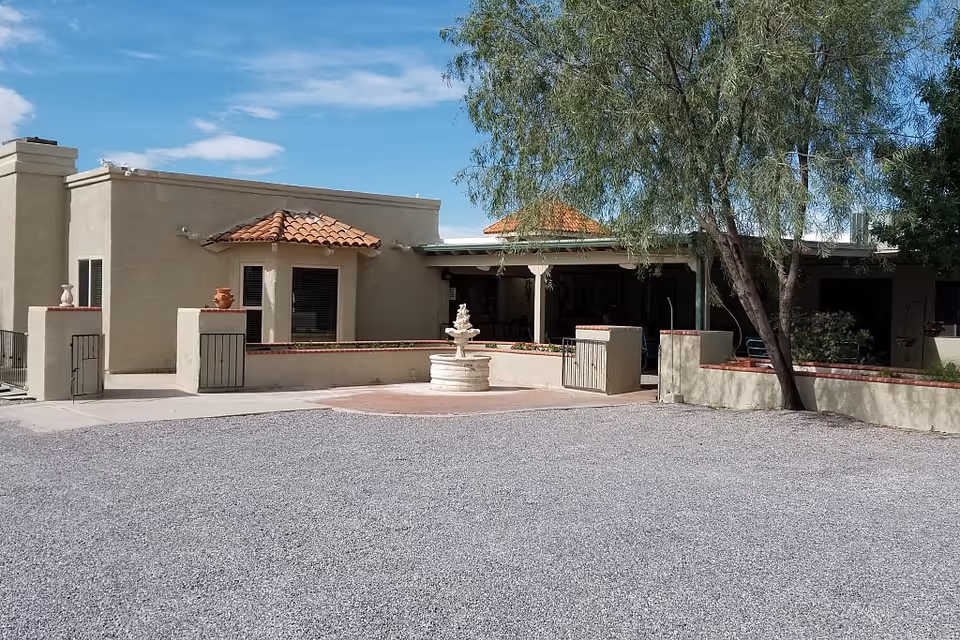 Exterior view of a single-story building with beige stucco walls and a red tile roof. There is a small courtyard area with a white stone fountain in the center, surrounded by low walls and gates. A tree is visible on the right side, and the ground is covered with gravel. The sky is partly cloudy with blue patches.