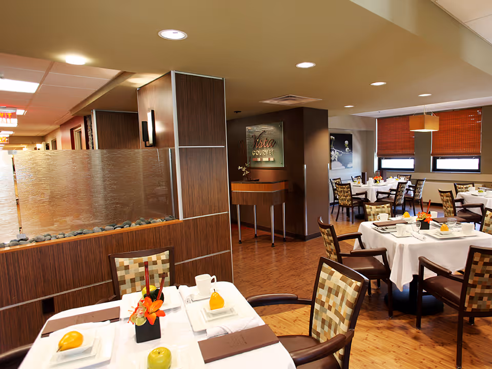 A dining room in Wexford Healthcare Center with multiple tables covered in white tablecloths, each set with plates, cups, napkins, and small decorative flower arrangements. The room features wooden flooring, patterned cushioned chairs, and windows with orange blinds. A water feature with rocks is visible near the entrance, and a sign reading 'Vista Gourmet' is mounted on a dark brown wall.