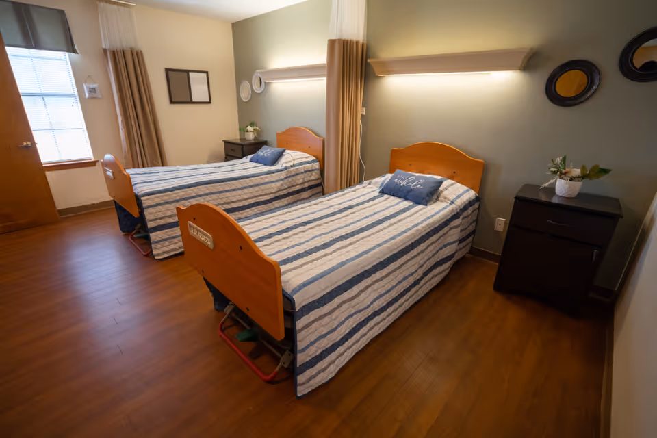 A bedroom in a senior living facility with two single beds, each with a striped blue and white bedspread and a decorative pillow. The room has wooden floors, a window with blinds and curtains, two nightstands with plants, and wall-mounted lights above each bed. There are also decorative mirrors and framed art on the walls.