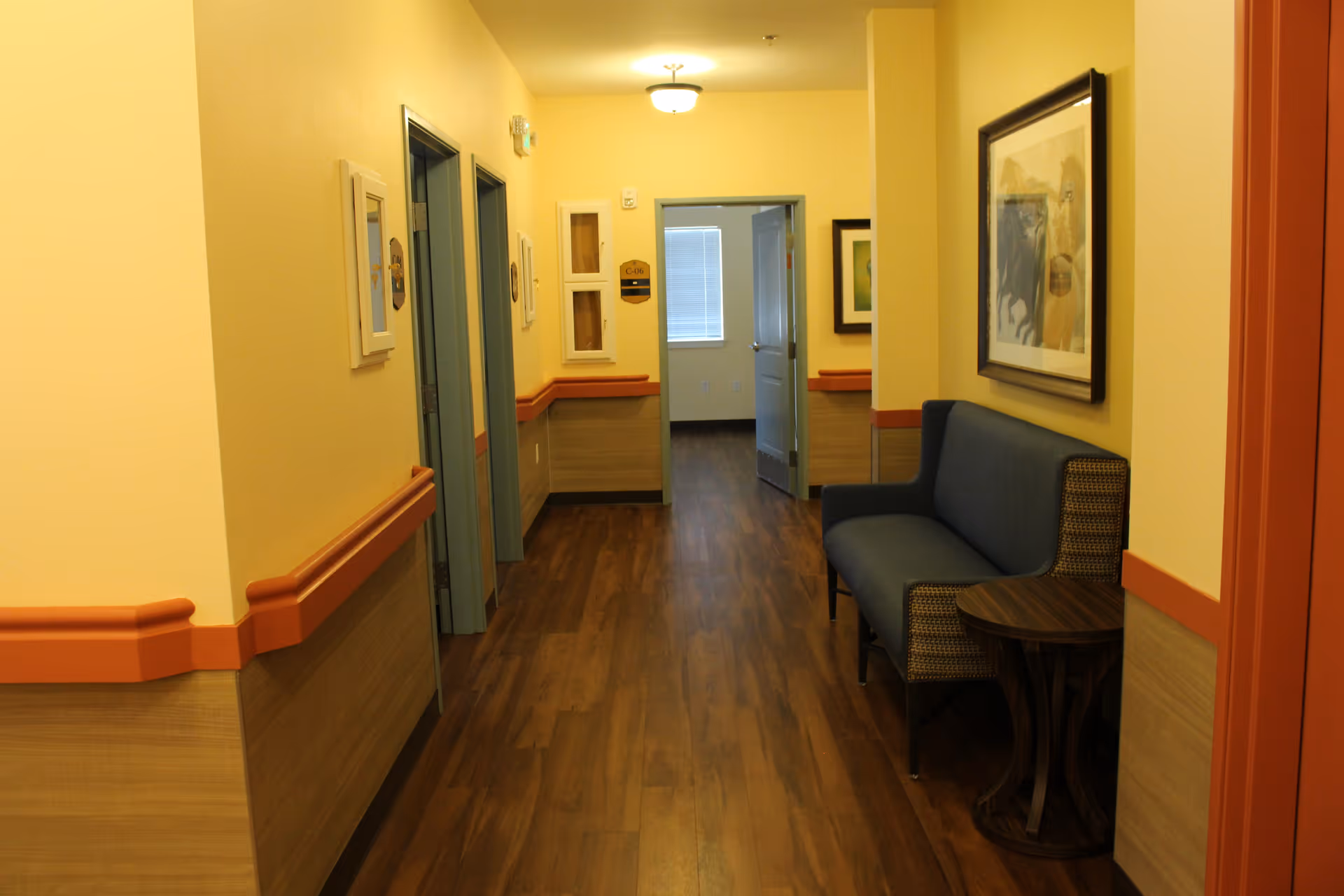 A well-lit hallway with wooden flooring and yellow walls featuring orange trim. On the right side, there is a blue cushioned bench with a small wooden side table next to it and a framed artwork hanging above. Several doorways are visible along the hallway, with one open door at the end revealing a room with a window.