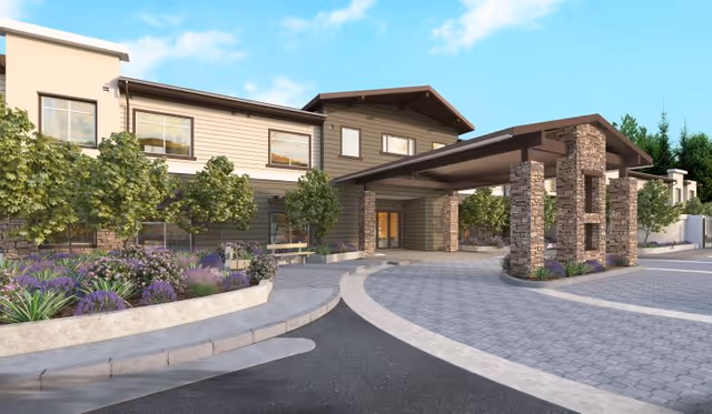 Exterior view of a senior living facility named Peninsula Pointe by Cogir, showing a two-story building with stone pillars supporting a covered entrance. The area is landscaped with trees, shrubs, and purple flowers under a blue sky with some clouds.