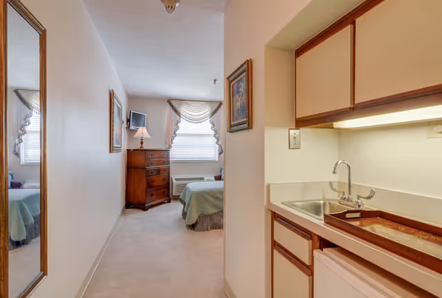 A studio-style senior living room with a kitchenette and sink on the right and a bed, dresser, and window at the far end.