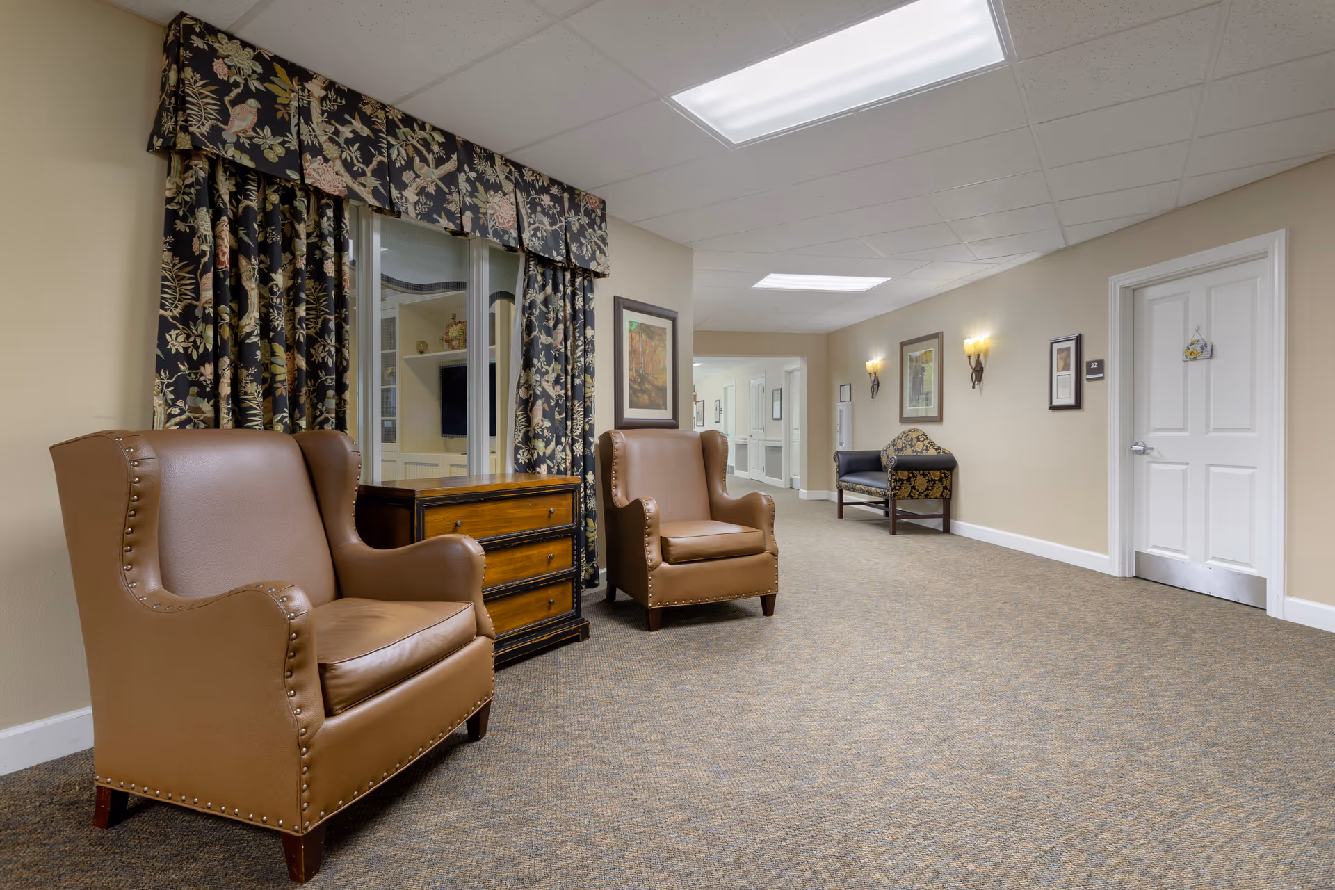 A carpeted hallway seating area with two brown leather armchairs, a wooden chest, patterned curtains and wall sconces.
