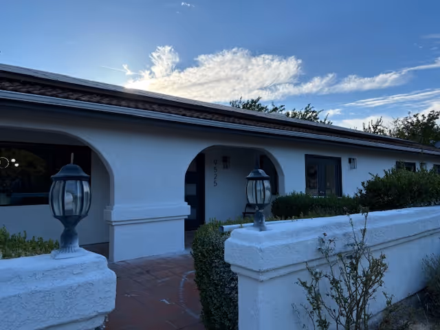 White stucco single-story building front with arched entryways, lantern-style lamps on a low perimeter wall, and shrubs under a blue sky.
