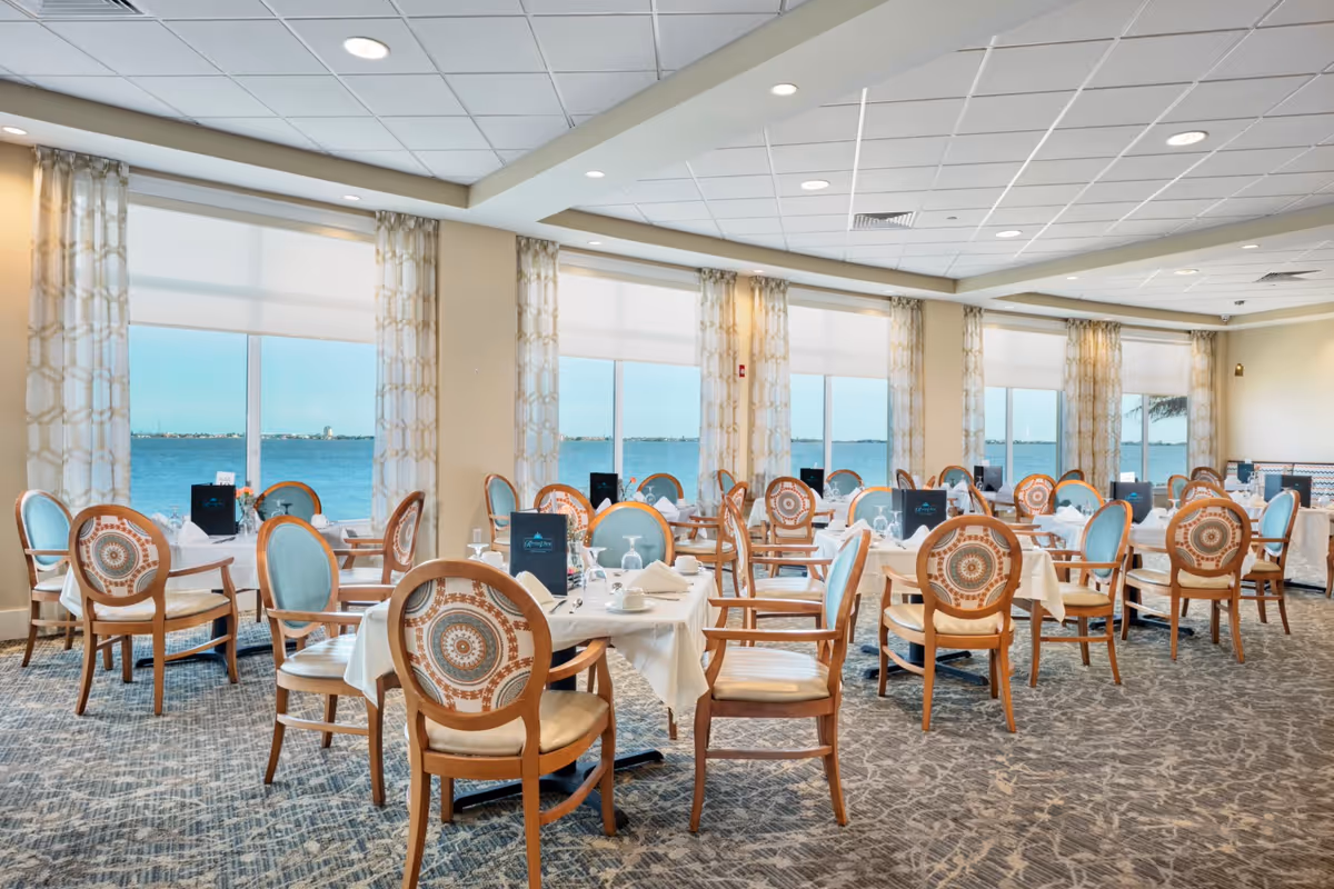 A bright dining room with multiple tables set with white tablecloths, napkins, cups, and glasses. The chairs have wooden frames with patterned backs and light blue cushions. Large windows with patterned curtains offer a view of a body of water outside.