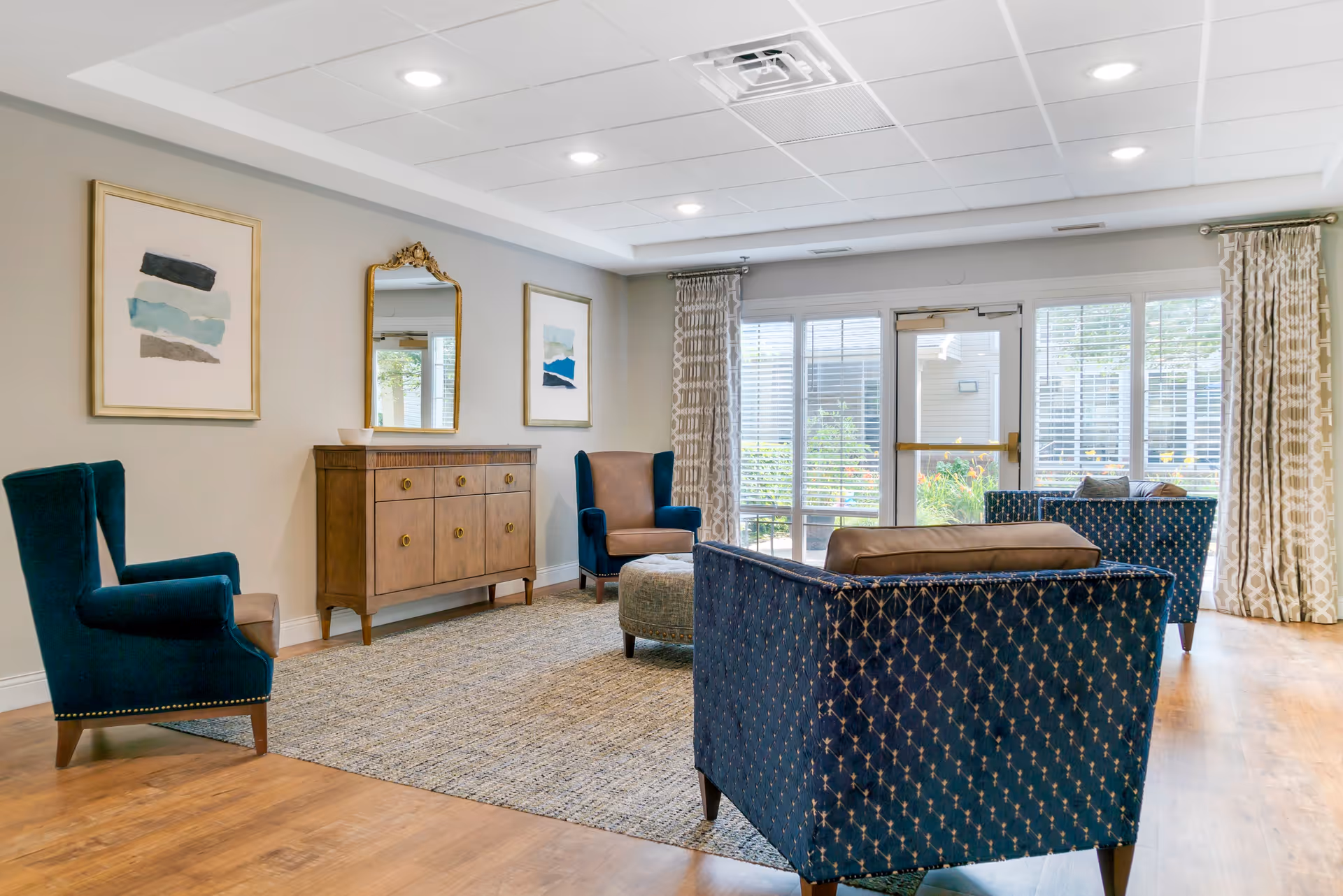 A bright and spacious living room area with wooden flooring, a large rug, and several upholstered chairs and a sofa in blue and brown tones. The room features a wooden sideboard with a decorative mirror and framed artwork on the wall. Large windows with white blinds and patterned curtains allow natural light to fill the space, and a glass door leads outside to a garden area.