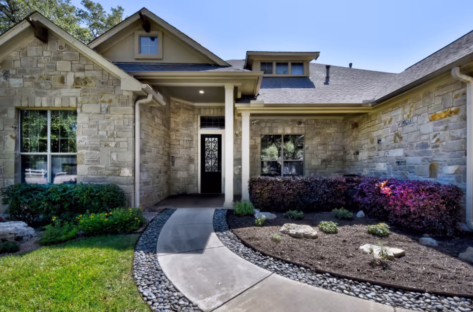 Front exterior view of a single-story stone house with a covered entrance, a black door with decorative glass, windows on either side, a curved concrete walkway bordered by small rocks, and landscaped garden beds with green shrubs and purple-leaved bushes under a clear blue sky.