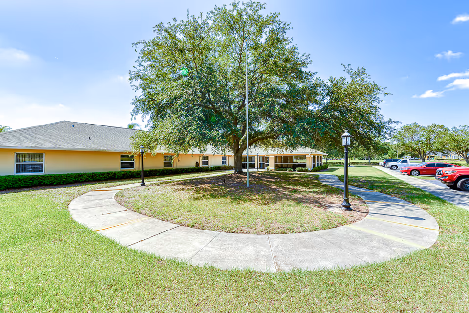 Low single-story senior living building with a large shade tree centered in a circular walkway and parked cars to the right.