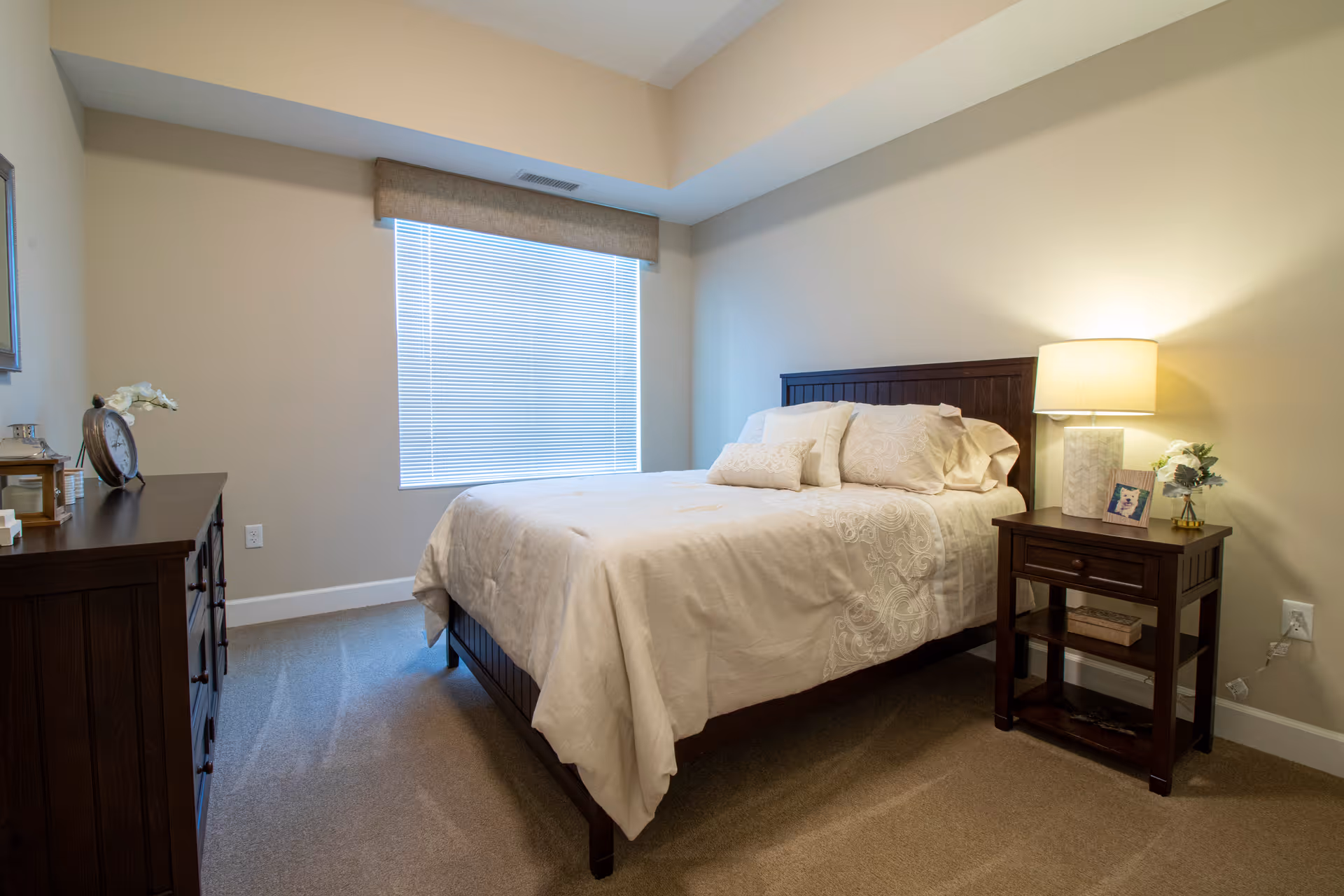 A neatly made bed with white bedding and multiple pillows in a bedroom. To the right of the bed is a wooden nightstand with a lamp, a small framed photo, and a small flower arrangement. On the left side of the room is a wooden dresser with decorative items including a clock and a small vase with flowers. The room has beige walls, a window with closed blinds, and carpeted flooring.
