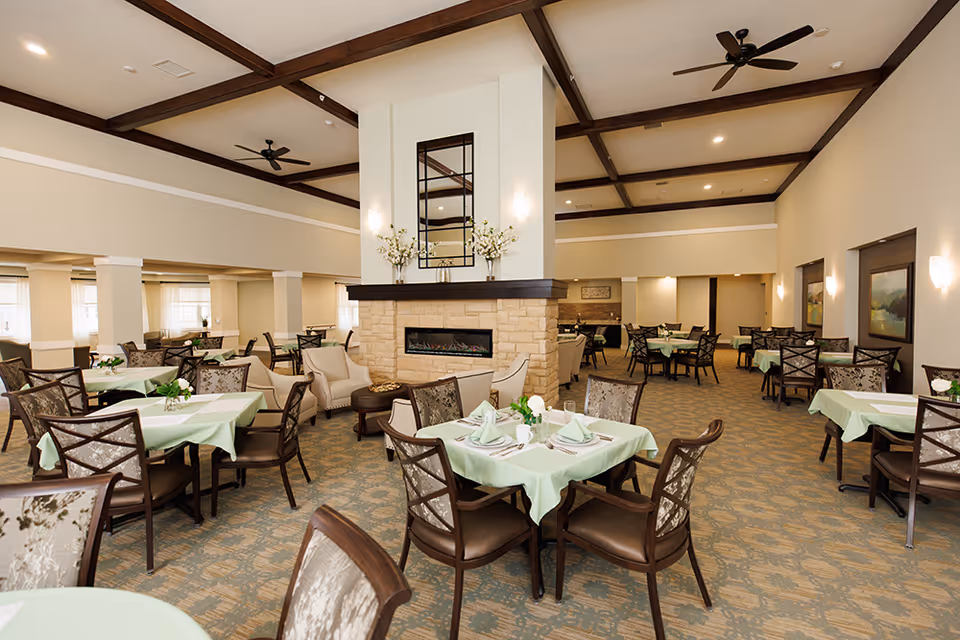 Spacious dining room with multiple tables covered in light green tablecloths, each set with napkins, plates, and glassware. The room features a central stone fireplace with a mirror and floral arrangements on the mantel. Comfortable chairs surround the tables, and the ceiling has exposed wooden beams with ceiling fans. Soft lighting and large windows provide a warm and inviting atmosphere.