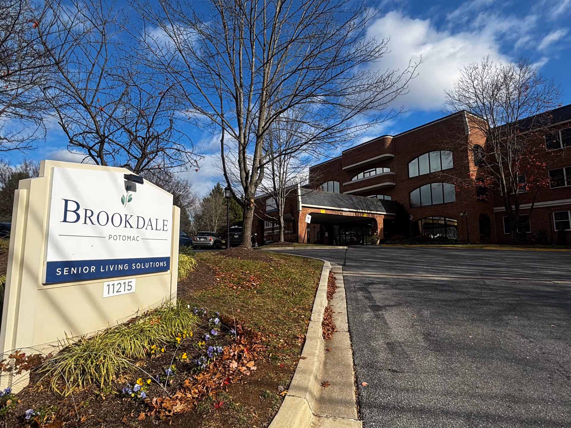 Exterior view of Brookdale Potomac senior living facility with a large sign in the foreground displaying the name and address. The building is a multi-story brick structure with large windows and a covered entrance. Trees without leaves and a partly cloudy blue sky are visible.