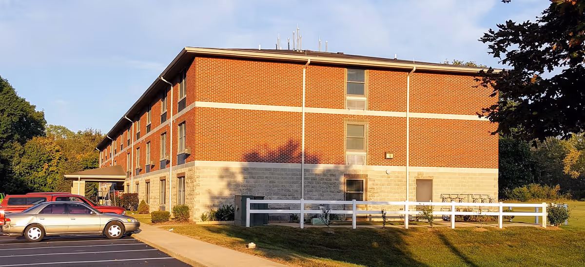 Exterior view of a three-story brick and stone apartment building with a parking lot in front and trees in the background, taken in daylight.
