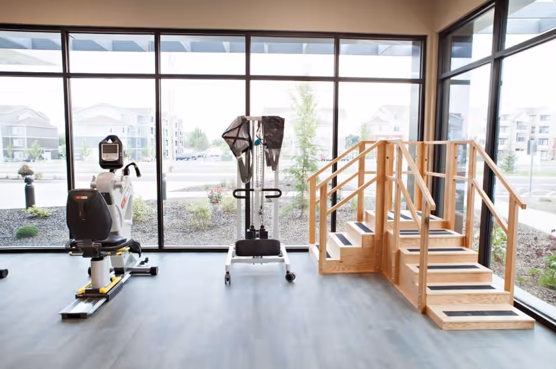 Bright therapy/exercise room with a recumbent exercise bike, a pulley machine, and wooden practice stairs in front of large floor-to-ceiling windows.