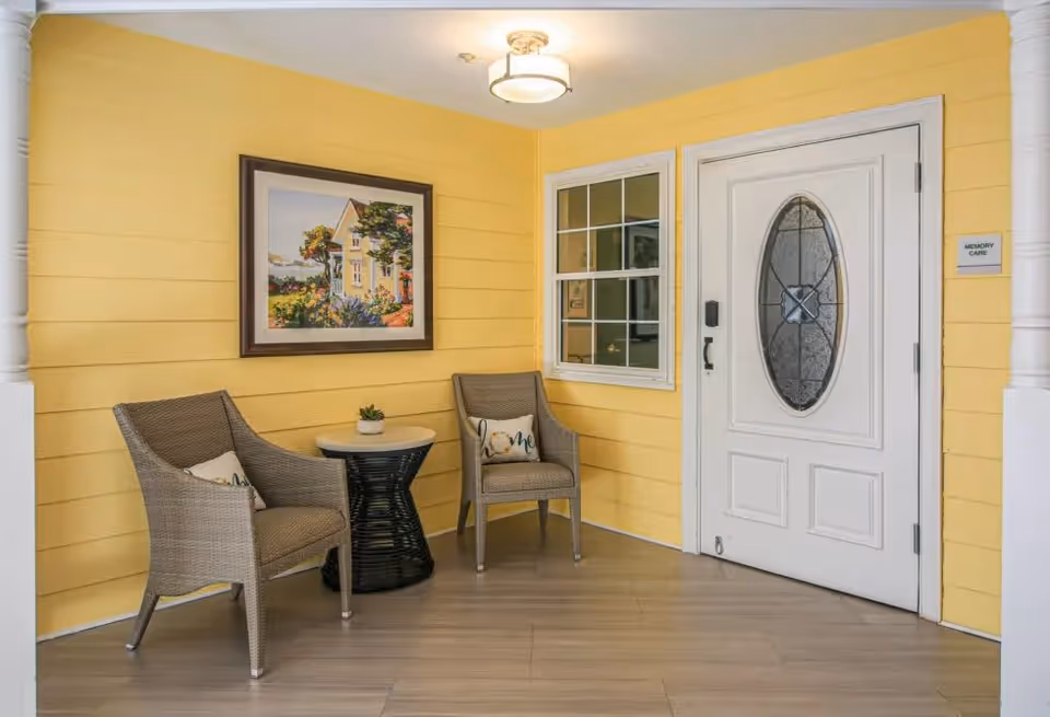 A cozy seating area with two wicker chairs and a small round table between them, placed against a yellow paneled wall. Above the chairs hangs a framed painting of a house with flowers. To the right, there is a white door with an oval glass window and a sign next to it that reads 'Memory Care'.