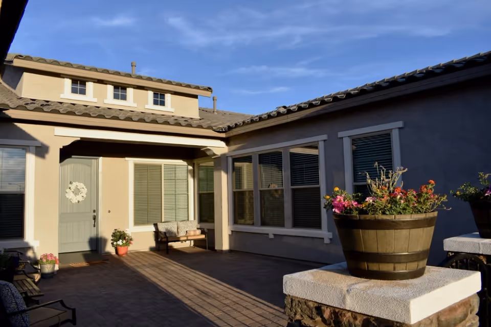 Sunlit courtyard of a single-story building showing a front door, windows, a bench, and potted flowers on stone pillars.