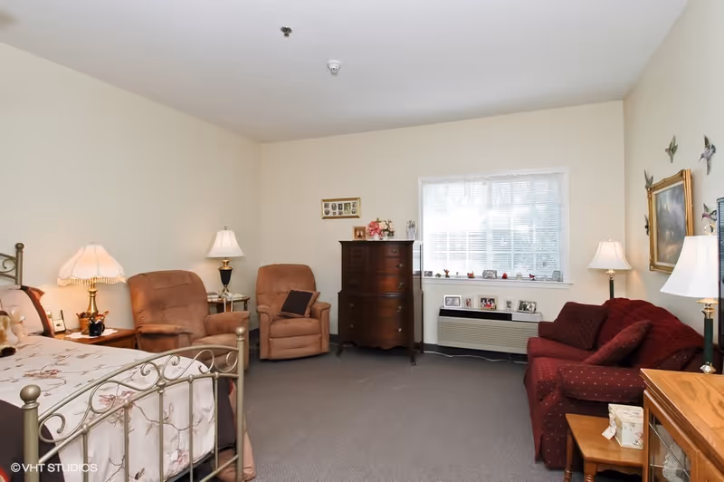 A cozy bedroom in a senior living facility featuring a metal-framed bed with floral bedding, two brown recliner chairs, a wooden chest of drawers, a red upholstered sofa with matching pillows, several table lamps, framed pictures on the walls, and a window with blinds letting in natural light.