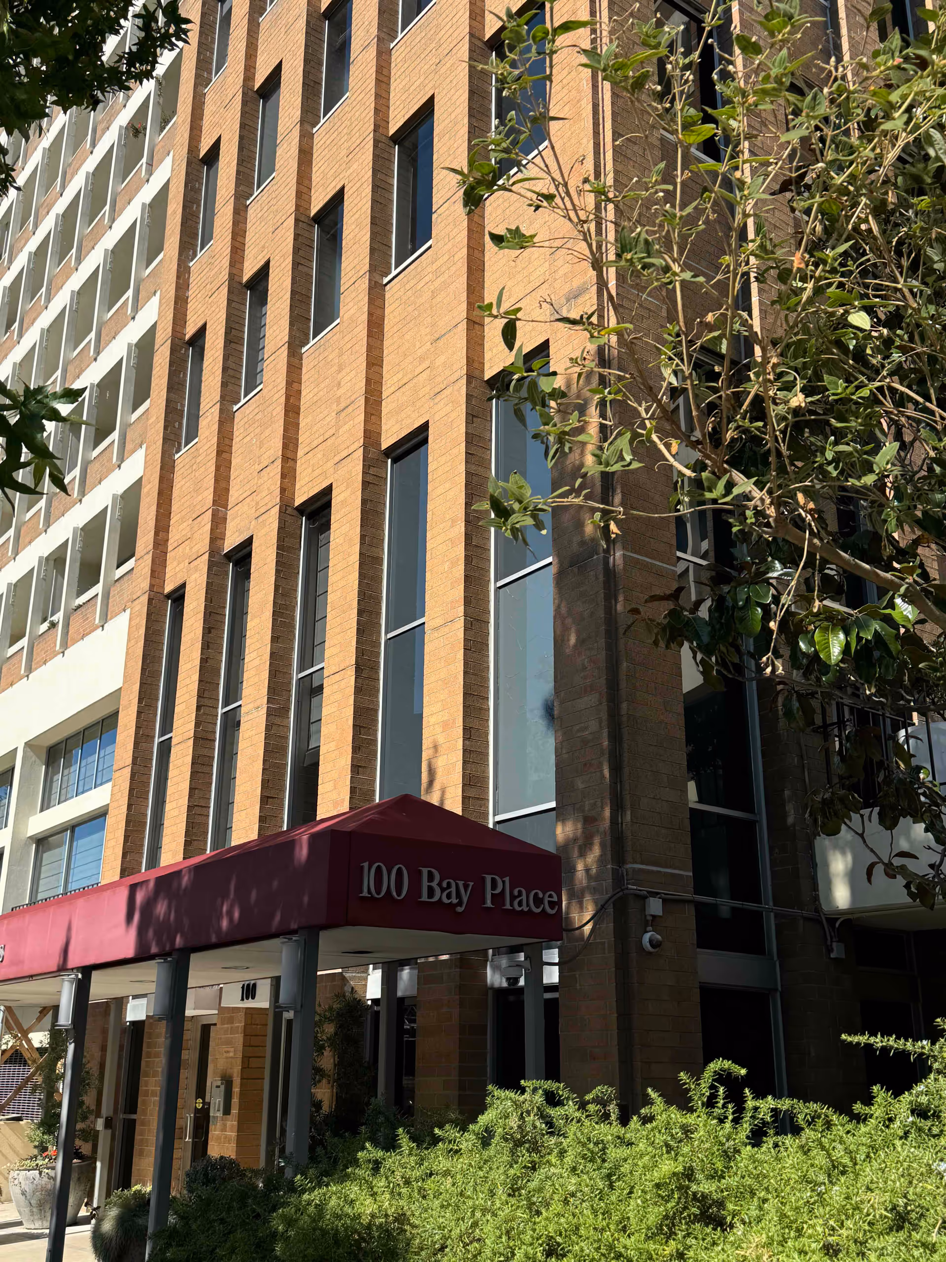 Exterior view of a multi-story brick building with large vertical windows and a maroon awning that reads '100 Bay Place'. Green shrubs and trees are visible in the foreground.