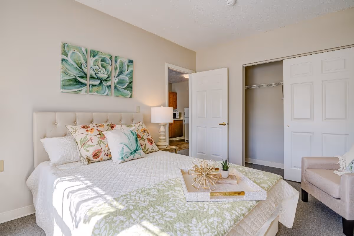 A bright bedroom with a neatly made bed featuring a white quilt and floral pillows. Above the bed is a three-panel green succulent artwork. To the right, there is an open closet with sliding doors and a beige armchair. A door leading to another room is open, showing part of a kitchen area with wooden cabinets and appliances. A bedside table with a lamp is next to the bed.