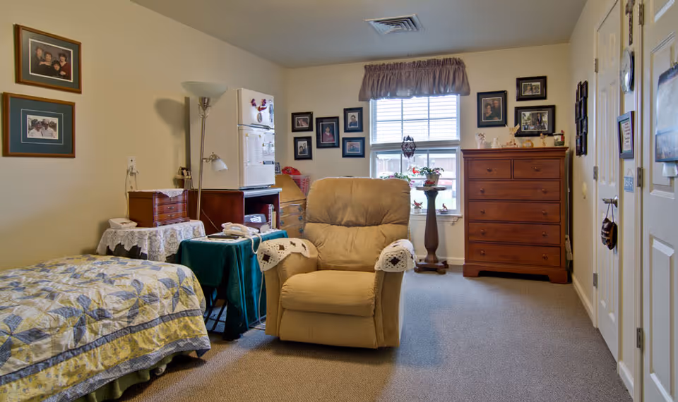 A cozy senior living bedroom with a single bed covered in a patterned quilt, a beige recliner chair with crocheted arm covers, a wooden dresser, a small table with a green cloth, a floor lamp, and several framed photos on the walls. A window with a valance lets in natural light, and there are decorative items on the dresser and windowsill.