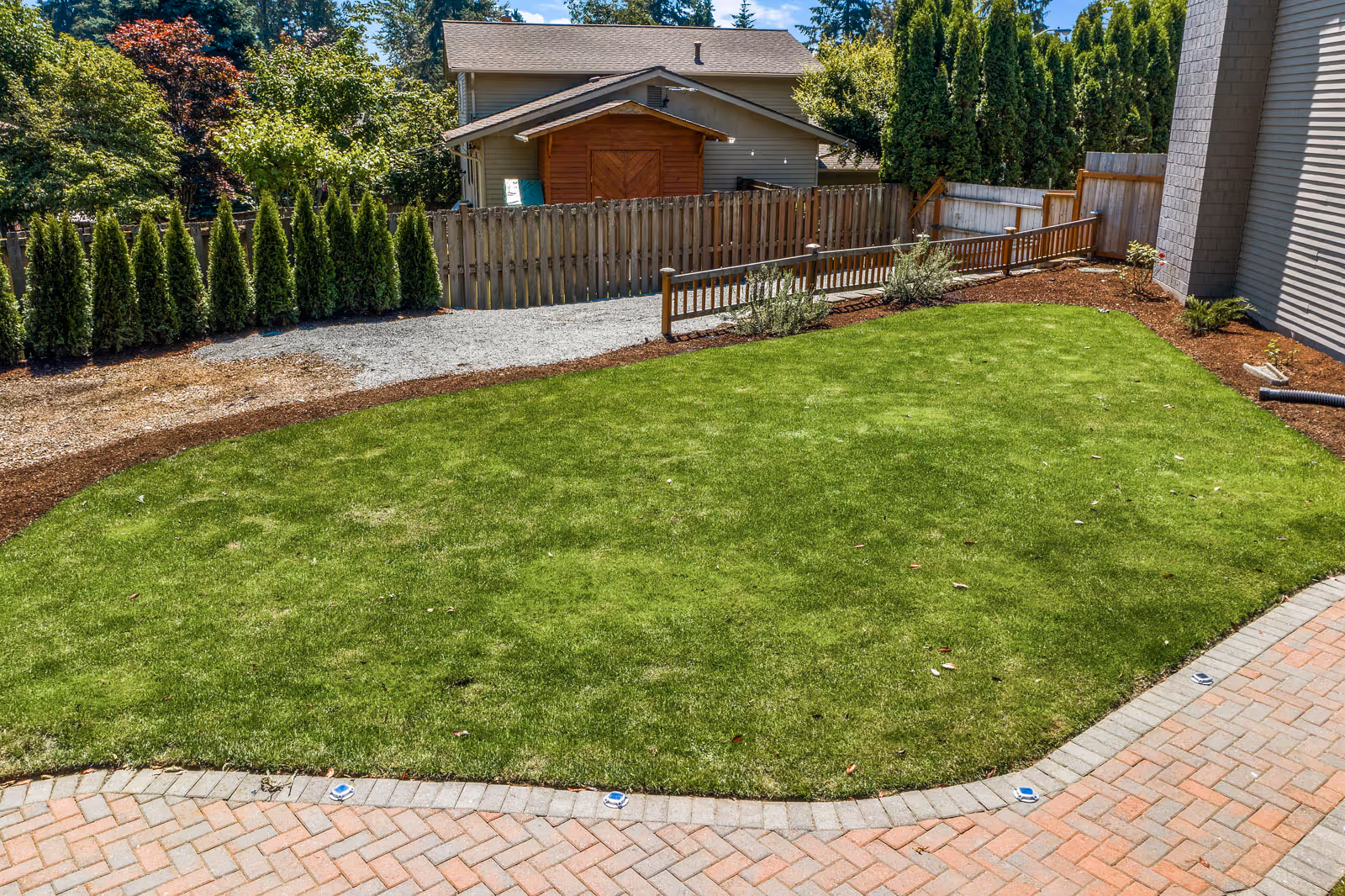 A well-maintained outdoor garden area with a green lawn bordered by a brick pathway. There are small shrubs and trees along the wooden fence, and a small wooden shed is visible in the background. The scene is sunny with clear skies.