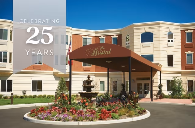 Exterior view of The Bristal Assisted Living at East Northport building with a covered entrance, a roundabout with a multi-tiered fountain, and a landscaped flower bed in front. The building has a mix of red brick and beige siding under a clear blue sky. Text overlay on the left side reads 'Celebrating 25 Years'.