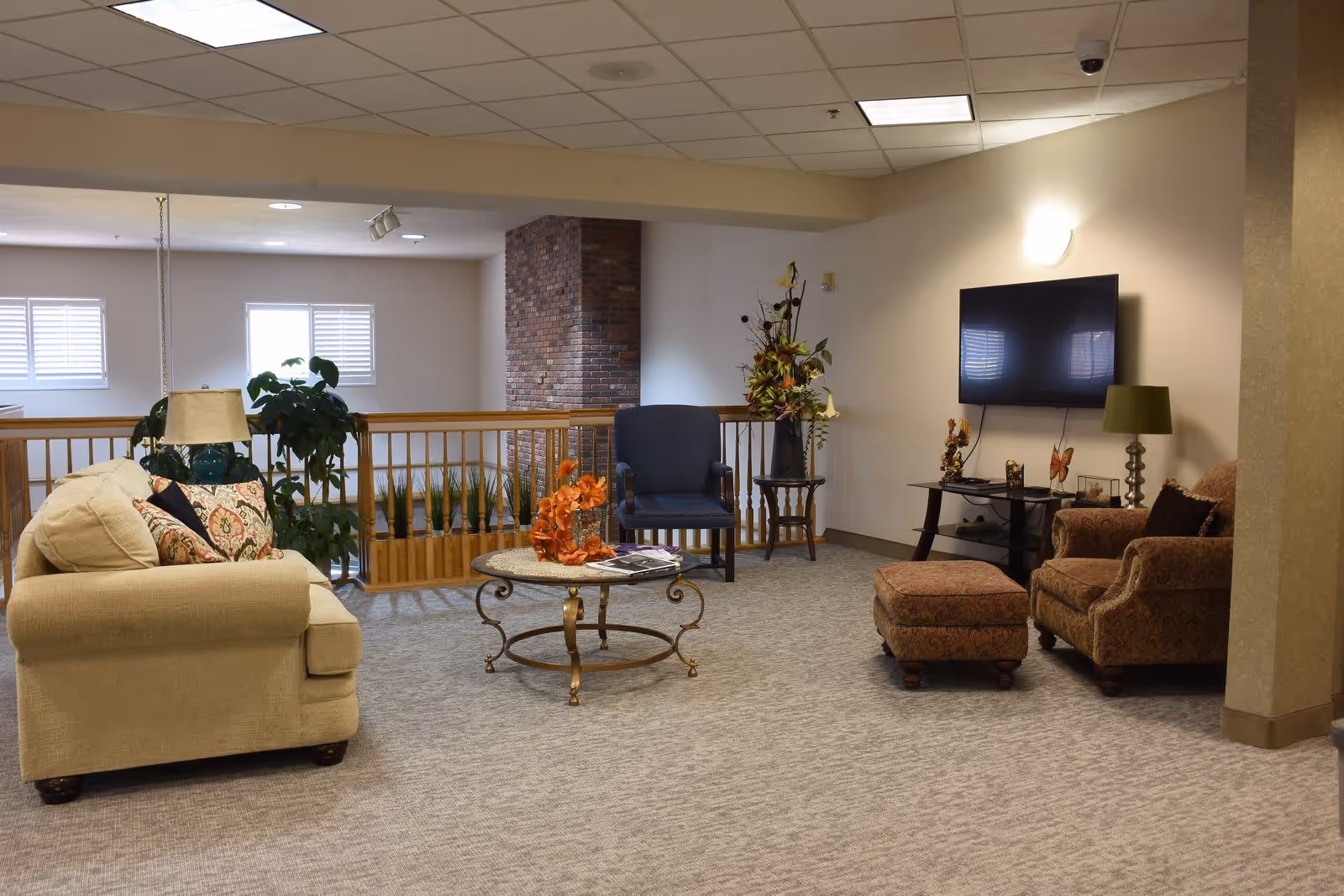 A cozy assisted living community lounge area with a beige sofa, a blue armchair, a patterned armchair with matching ottoman, a round glass coffee table with decorative items, a flat-screen TV mounted on the wall, and various plants and floral arrangements. The space has carpeted flooring and a ceiling with recessed lighting.