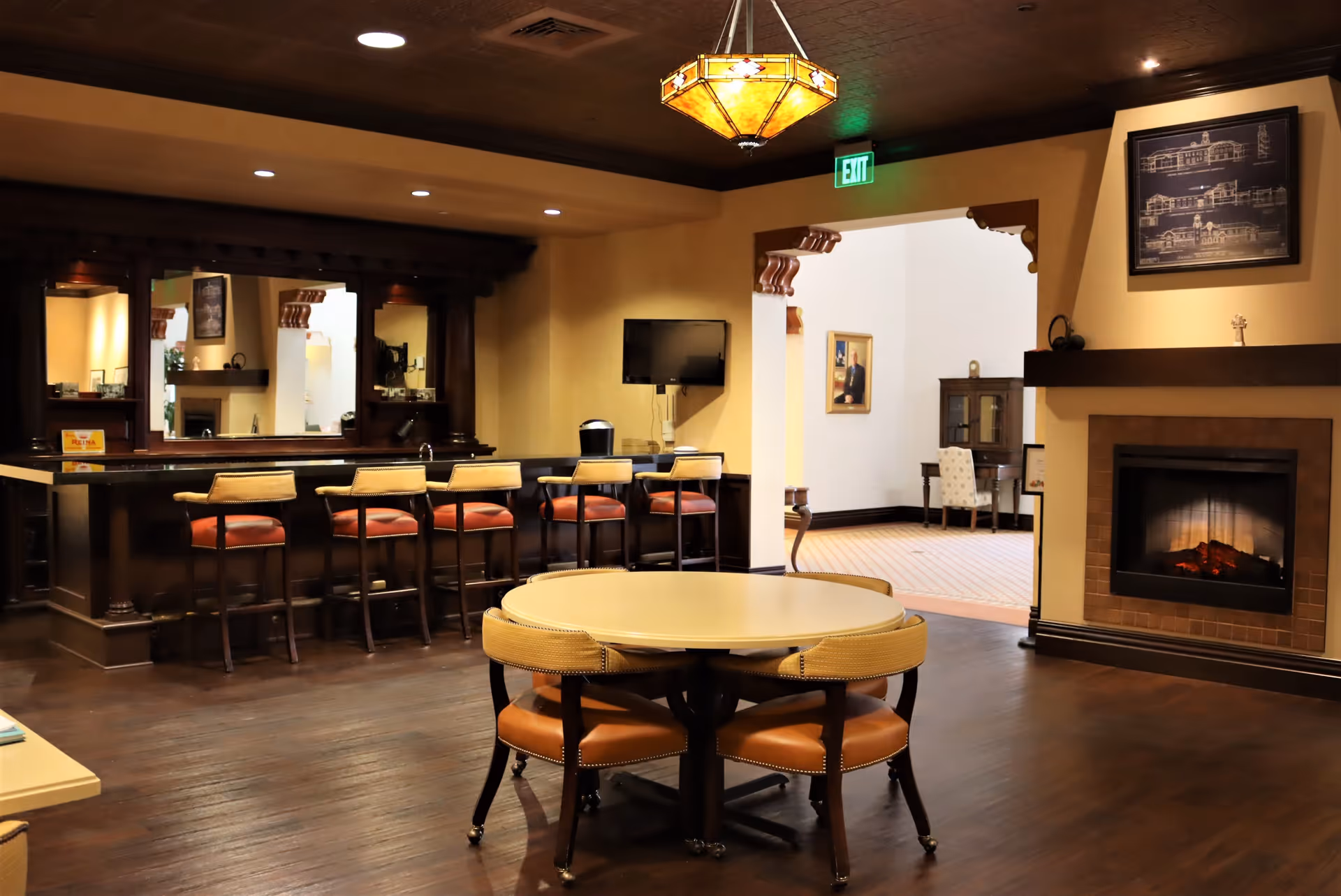A cozy interior room with a round table surrounded by four cushioned chairs in the foreground. To the left, there is a bar area with five high chairs and a large mirror behind the bar. On the right side, there is a fireplace with a framed blueprint above it. The room has warm lighting and wooden flooring, with an exit sign above a doorway leading to another room with a desk and chair.