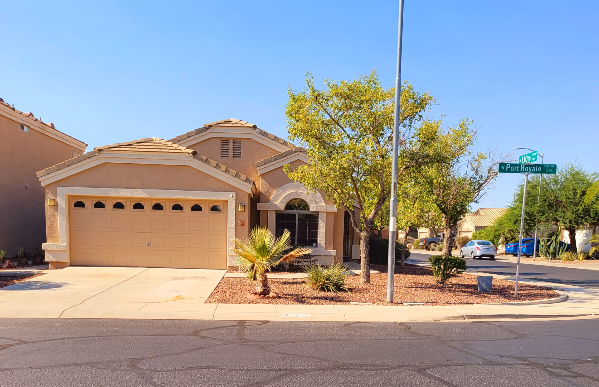 Exterior view of a single-story house with a two-car garage, a driveway, and a landscaped front yard featuring trees and shrubs. The house is located on a corner lot with street signs visible for W Port Royale and 1300 E. The sky is clear and blue.